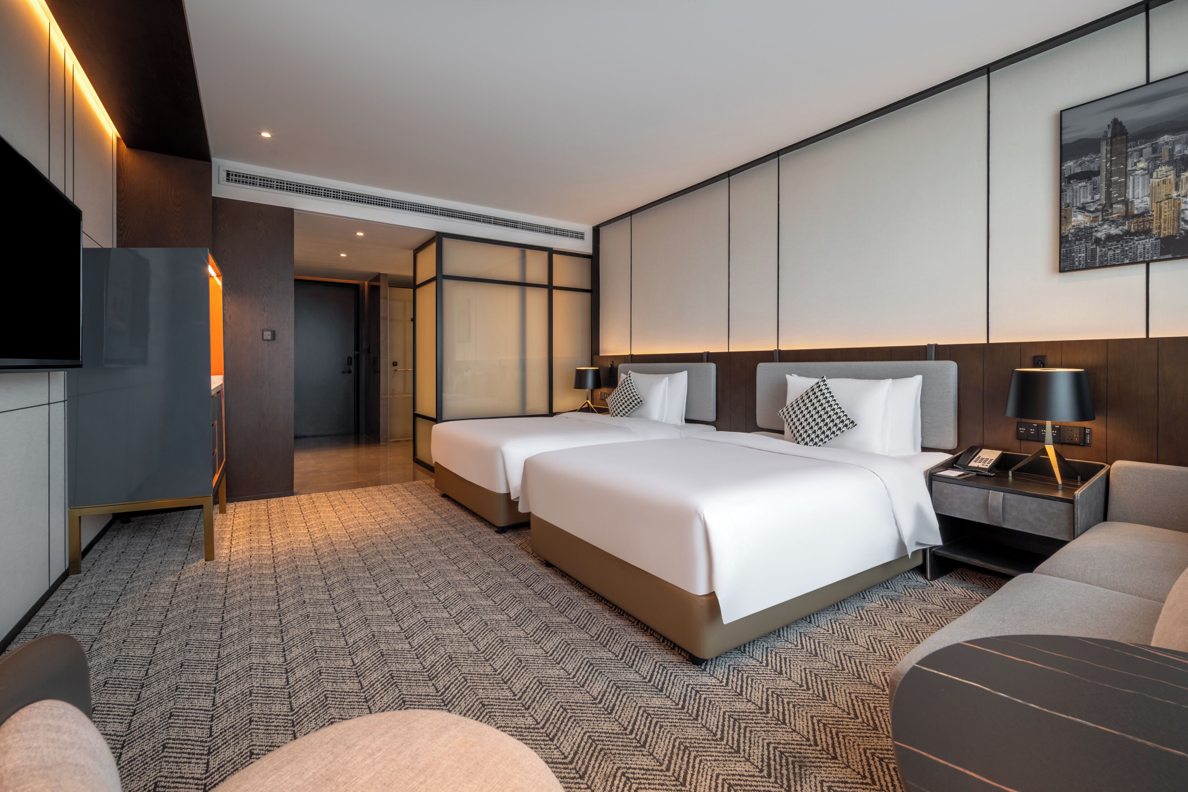 A hotel room with two beds, a TV, a desk with a lamp, and a cityscape view.