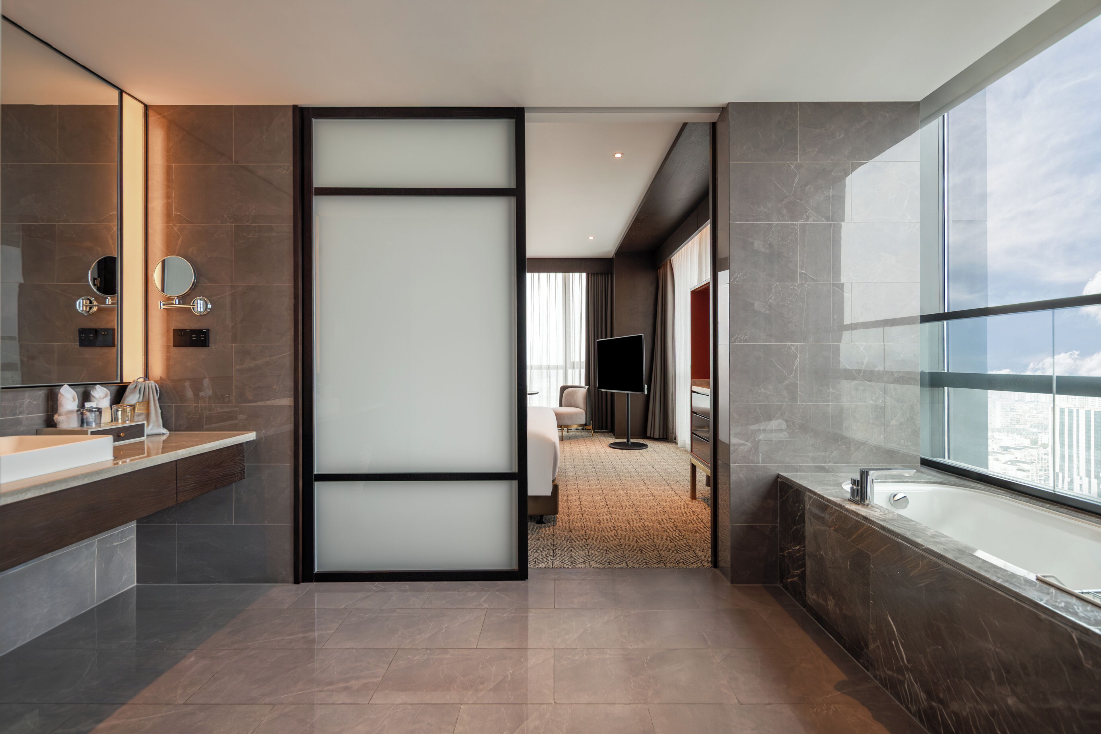 A modern hotel bathroom with a large bathtub, a vanity with a mirror, and a television in the background.