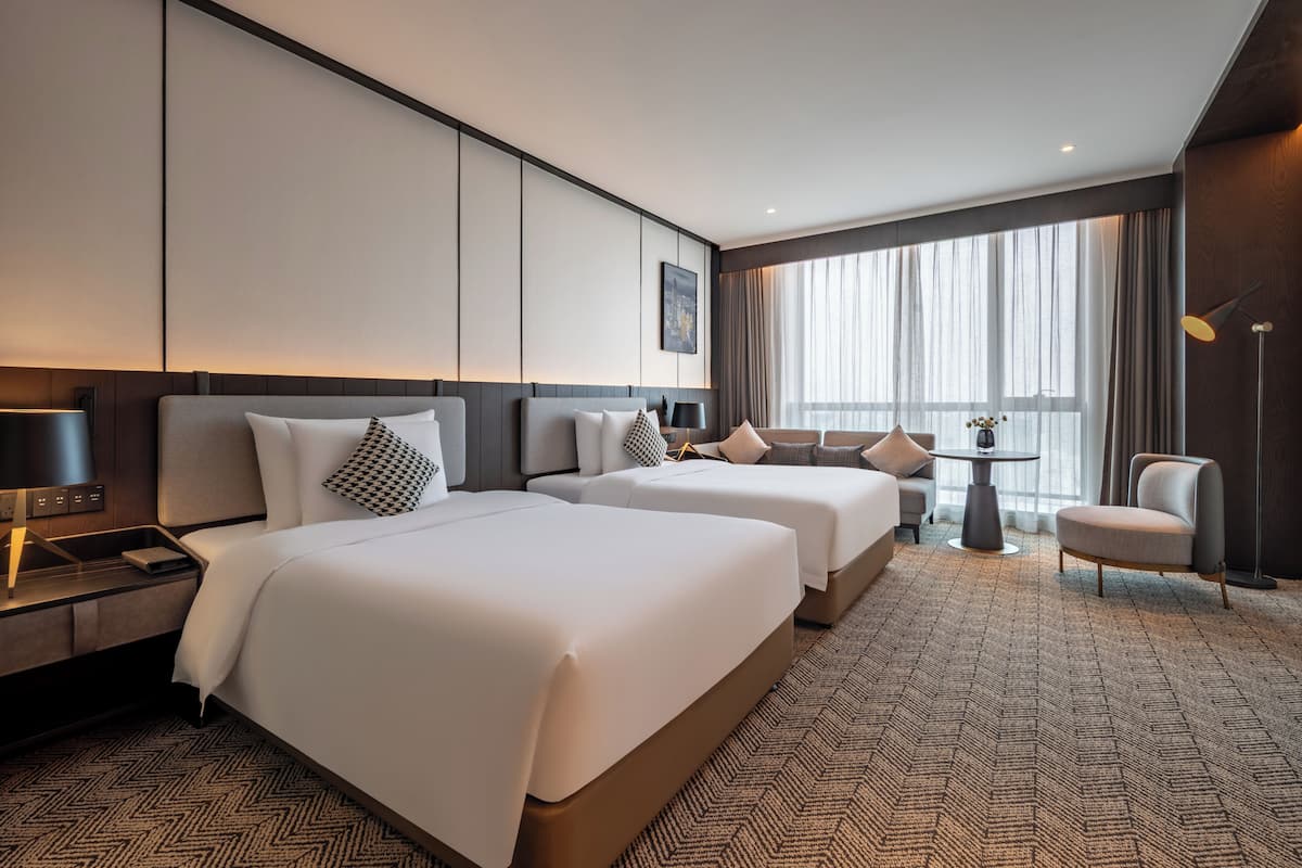 A hotel room with two beds, a desk, a chair, a table, and a large window with curtains.