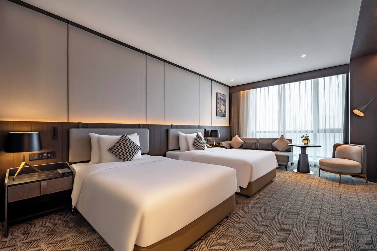 A hotel room with two beds, a desk, a chair, a small table, and a large window with curtains.