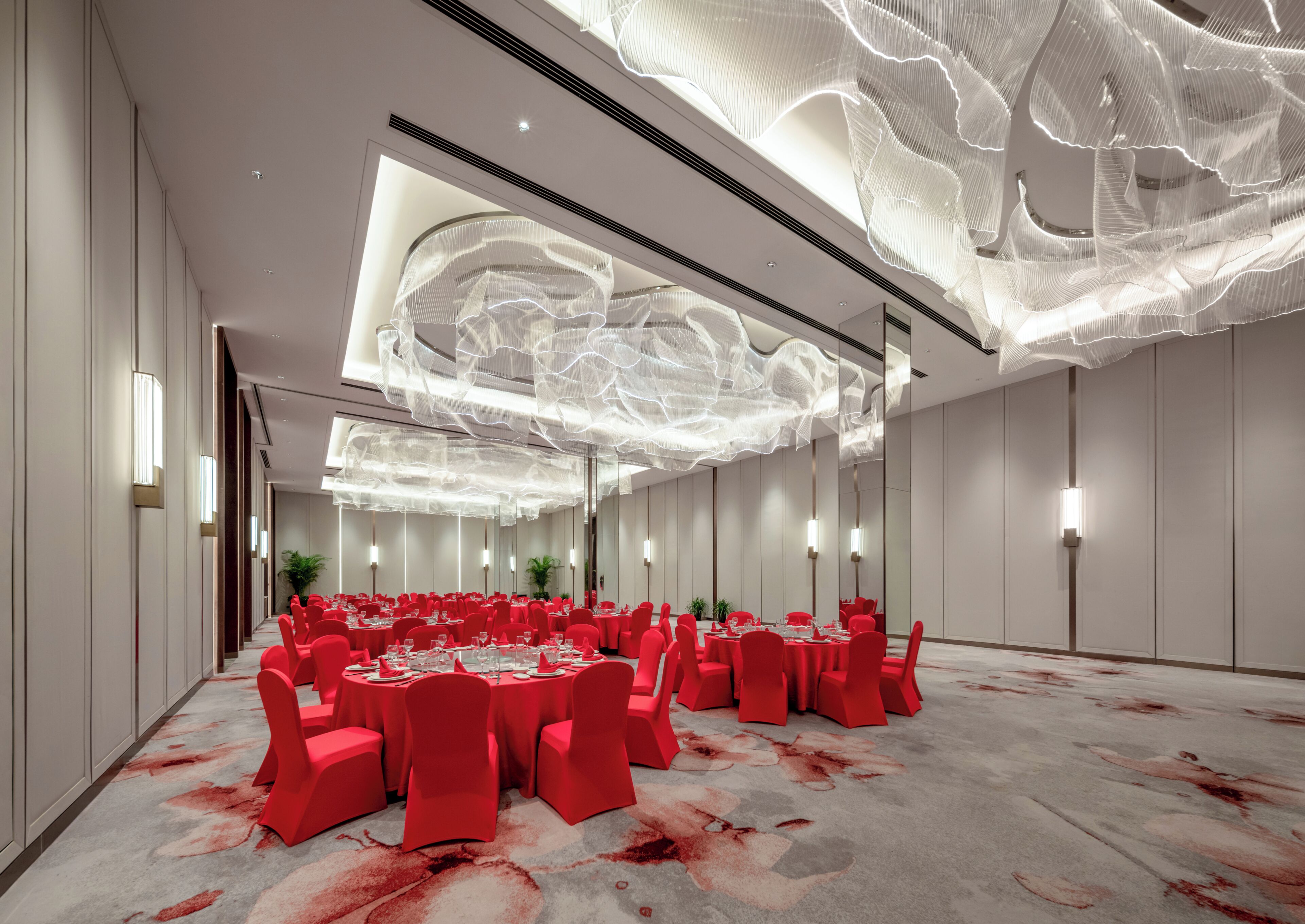 A spacious banquet hall with round tables, red chairs, and a modern ceiling design.