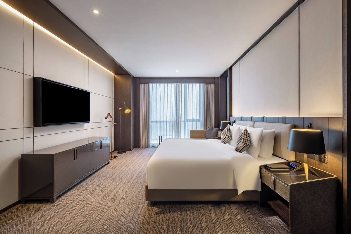A modern hotel room with a large bed, a flat-screen TV, a bedside table with a lamp, and a view of the city through large windows.