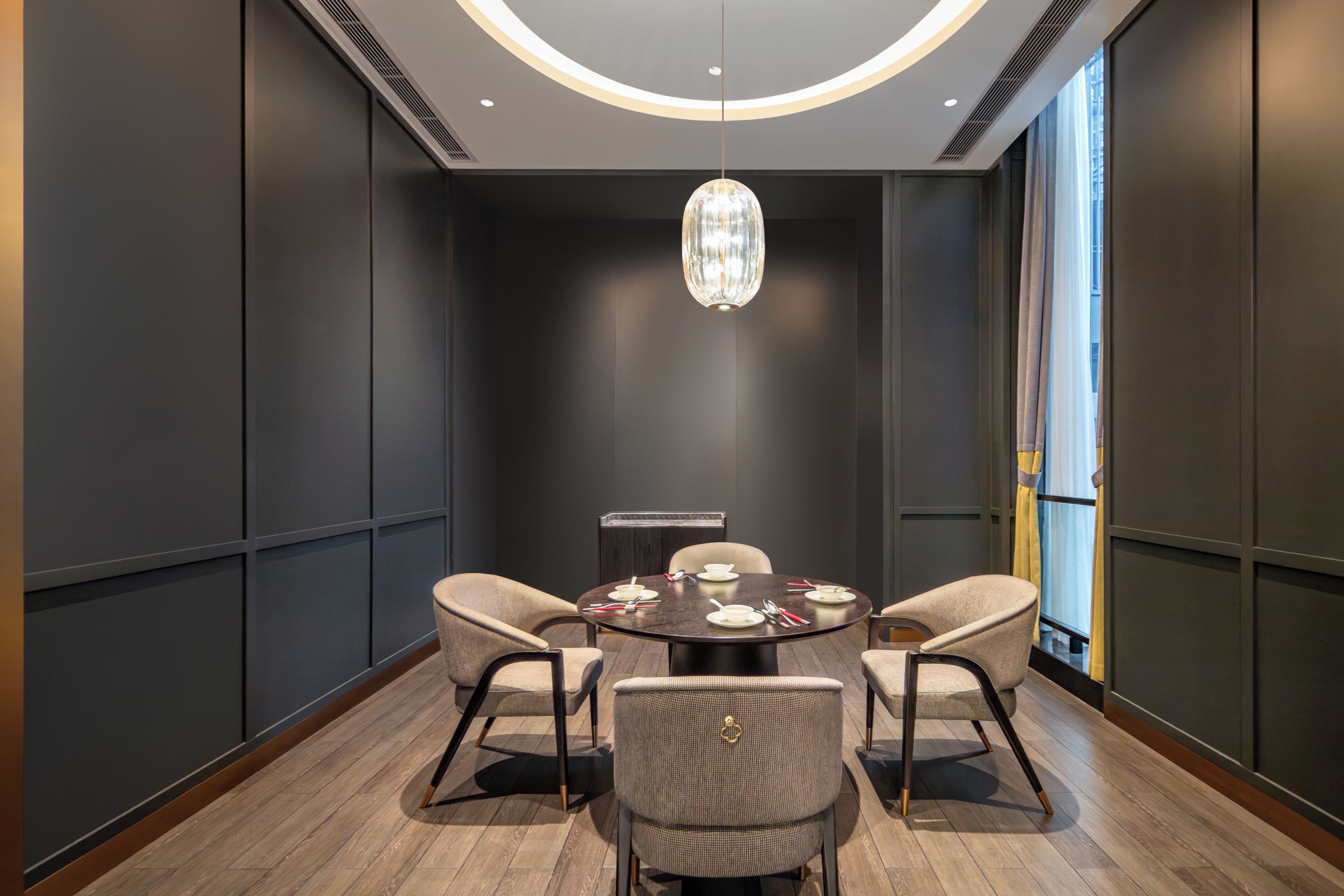 A conference room with a round table, four chairs, a chandelier, and a large window with curtains.