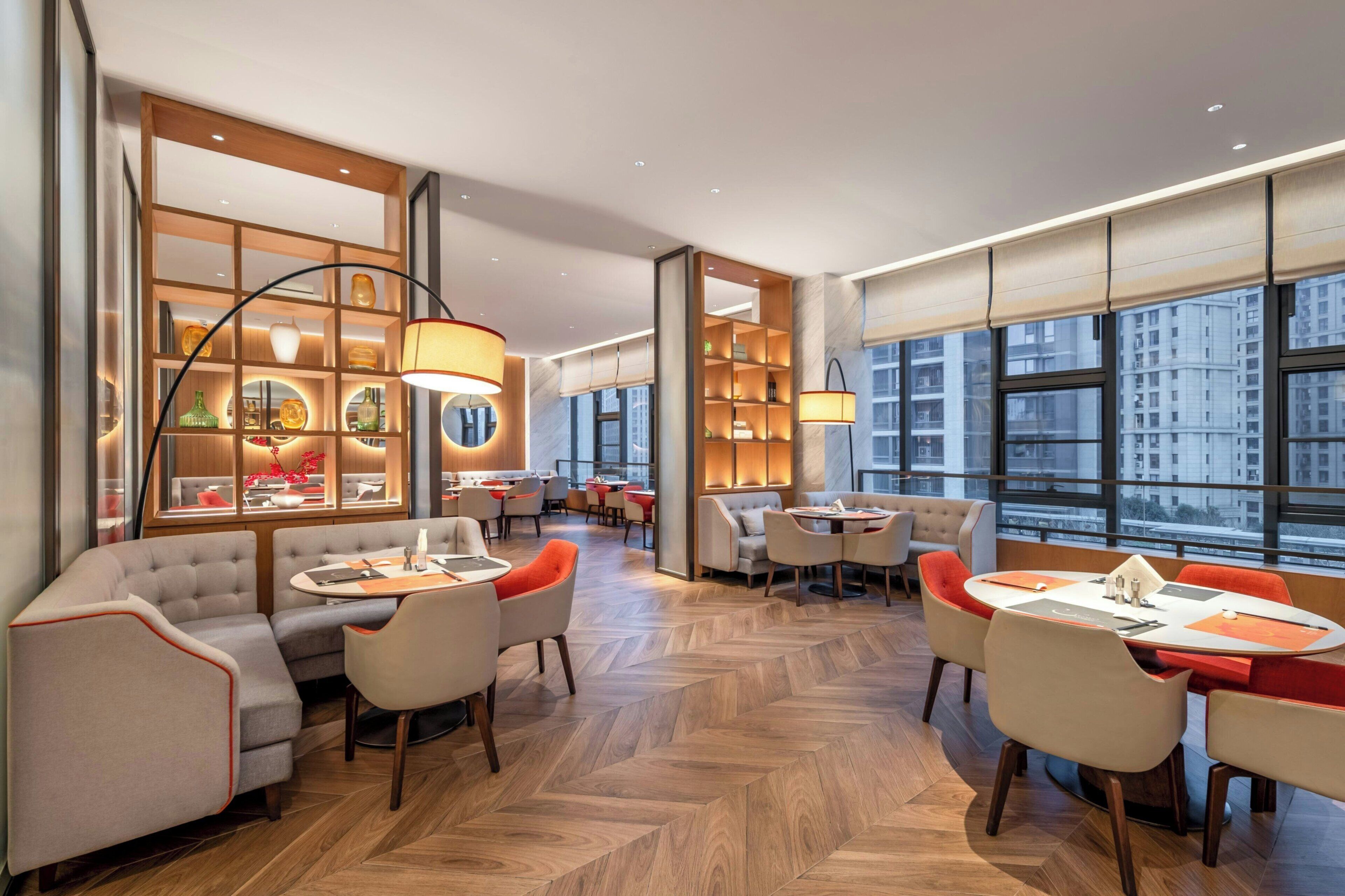 Un moderno vestíbulo de hotel con suelo de madera en patrón de espiga, zonas de estar con mesas y sillas, y amplias ventanas con vistas a la ciudad.