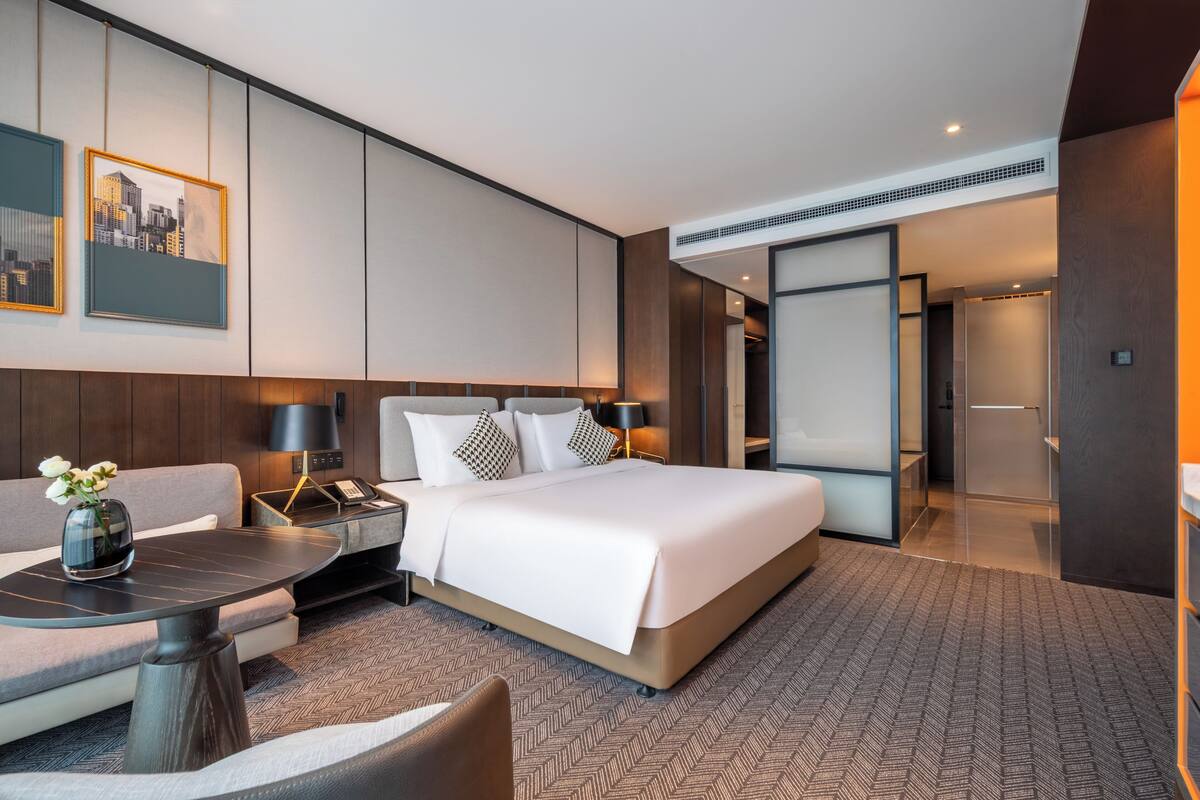 A modern hotel room with a large bed, a sofa, a desk, and a cityscape painting on the wall.