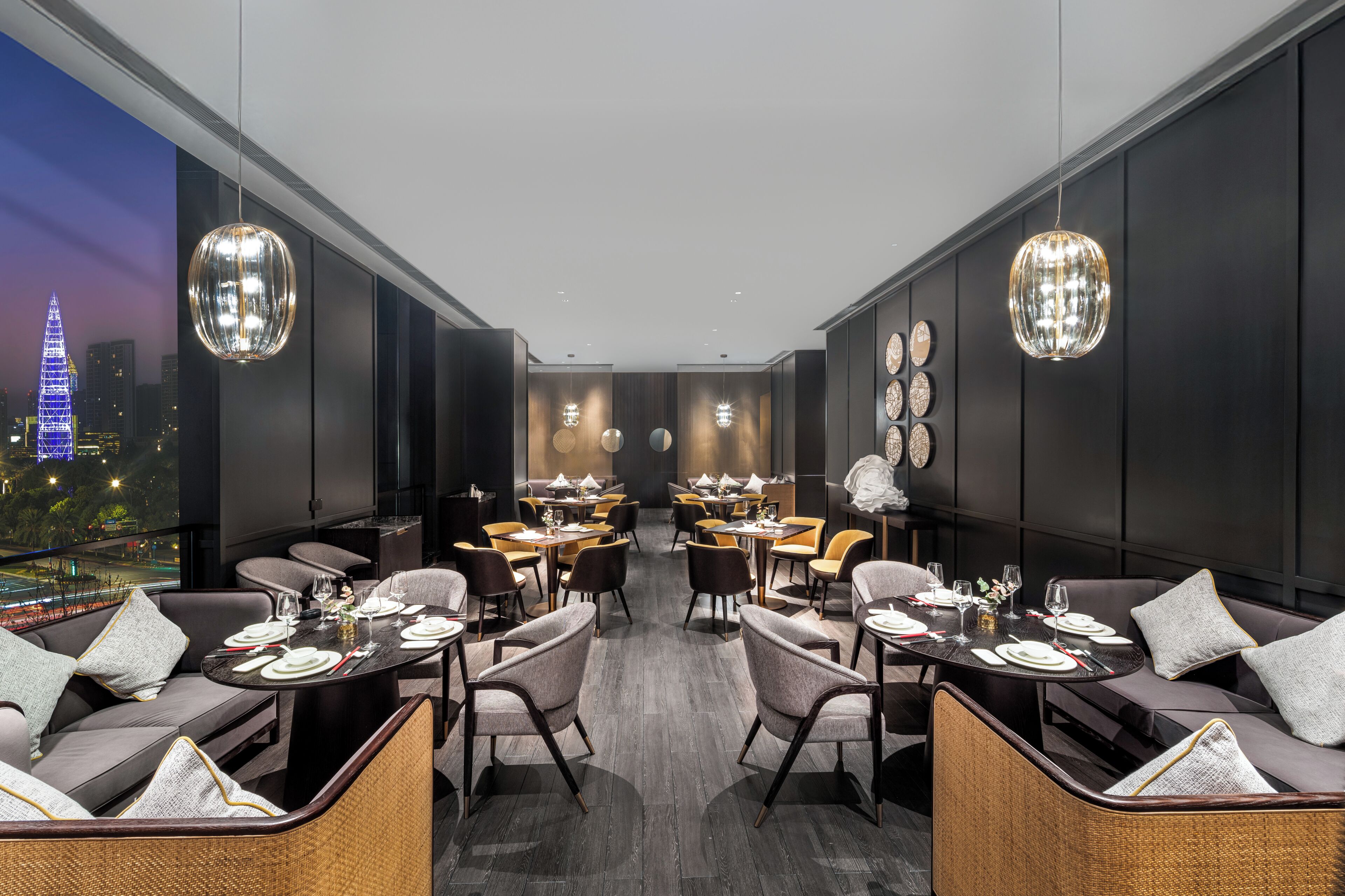 En moderne restaurant med høje lofter, mørke vægge og store vinduer med udsigt over byen. Bordene er dækket med hvide duge, og der er flere stole.