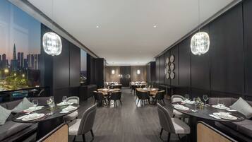Un espace de restauration moderne doté de plusieurs tables et chaises, offrant une vue sur la ville grâce à de grandes fenêtres, et agrémenté d’œuvres d’art décoratives sur les murs.