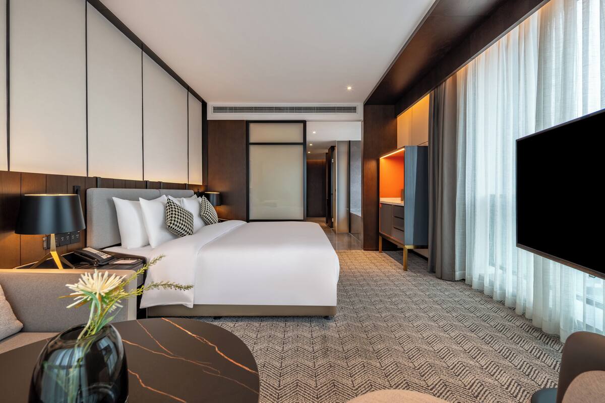 A modern hotel room with a large bed, a flat-screen TV, a desk with a lamp, and a view of the city through large windows.