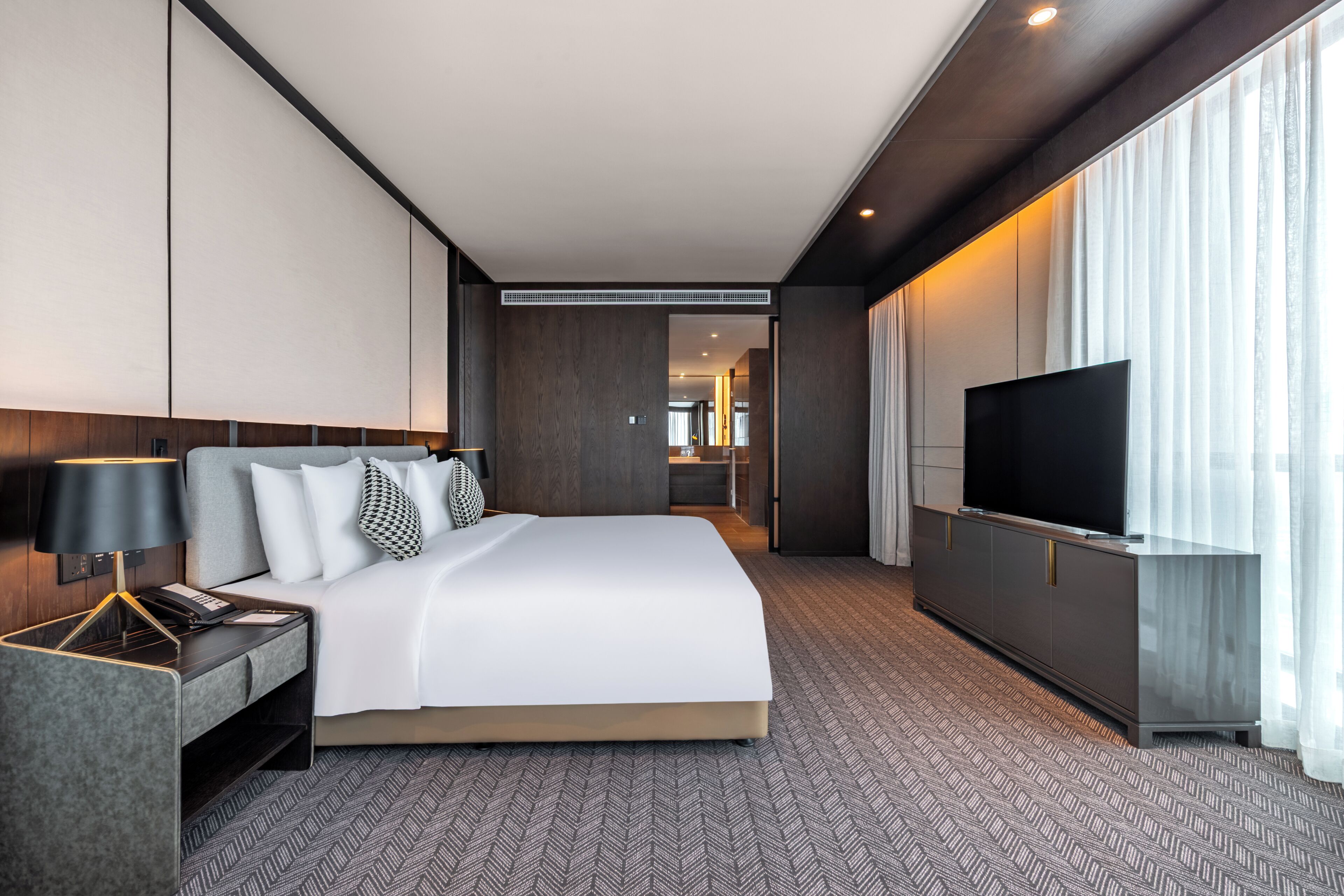 A modern hotel room with a large bed, a TV, a desk with a lamp, and a view of the city through the window.