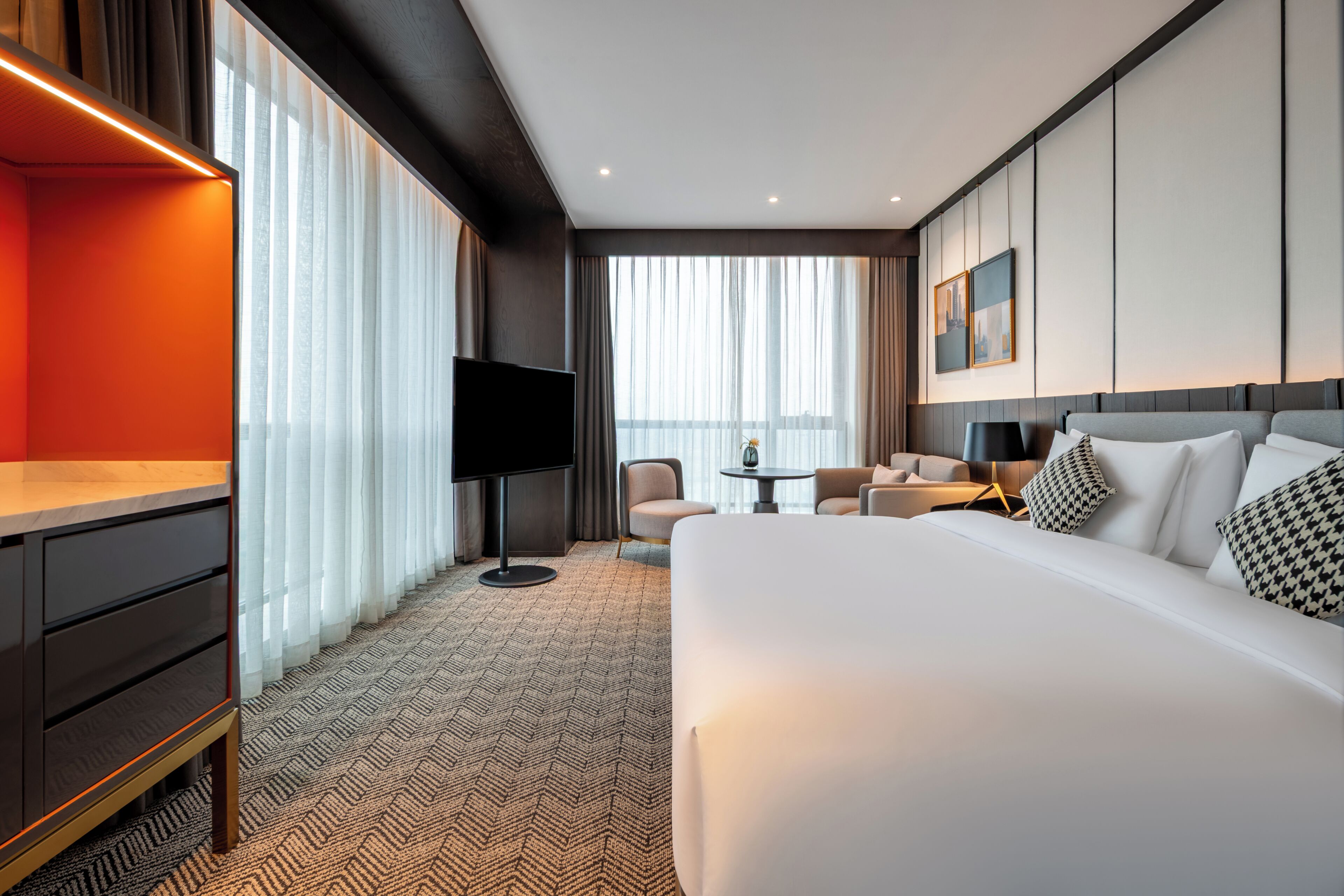 Quarto de hotel com uma cama grande, uma escrivaninha, uma TV, uma cadeira, uma mesa pequena e uma janela com cortinas.