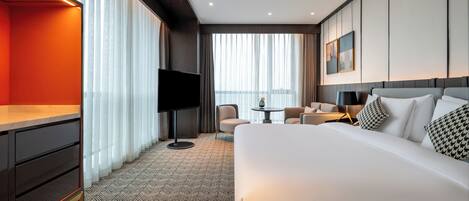 Quarto de hotel com uma cama grande, uma escrivaninha, uma TV, uma cadeira, uma mesa pequena e uma janela com cortinas.