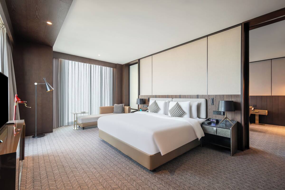 A modern hotel room with a large bed, bedside tables, a TV, and a large window with curtains.