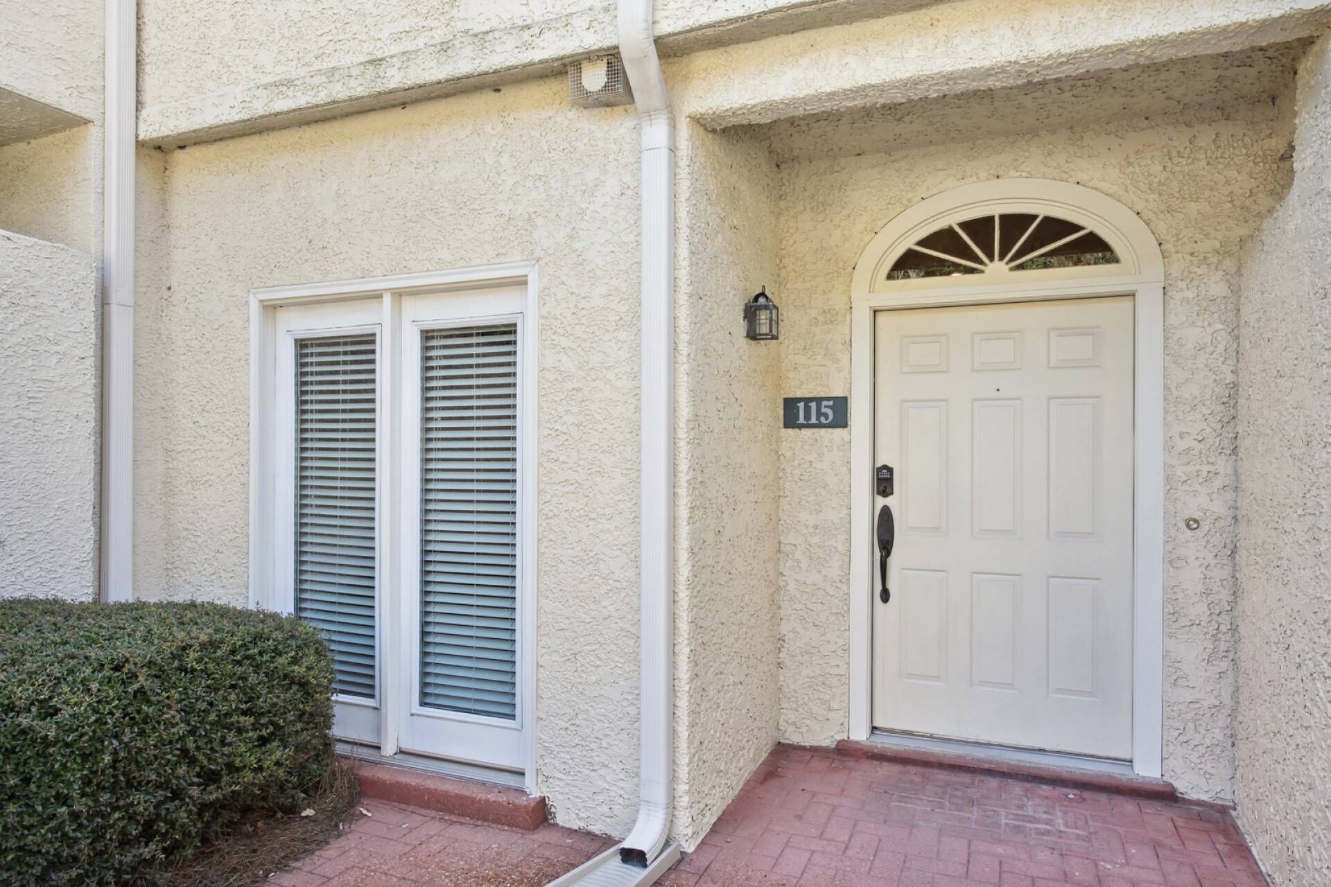 A building entrance with a white door marked with the number 115, a white mailbox, and a small bush to the left.