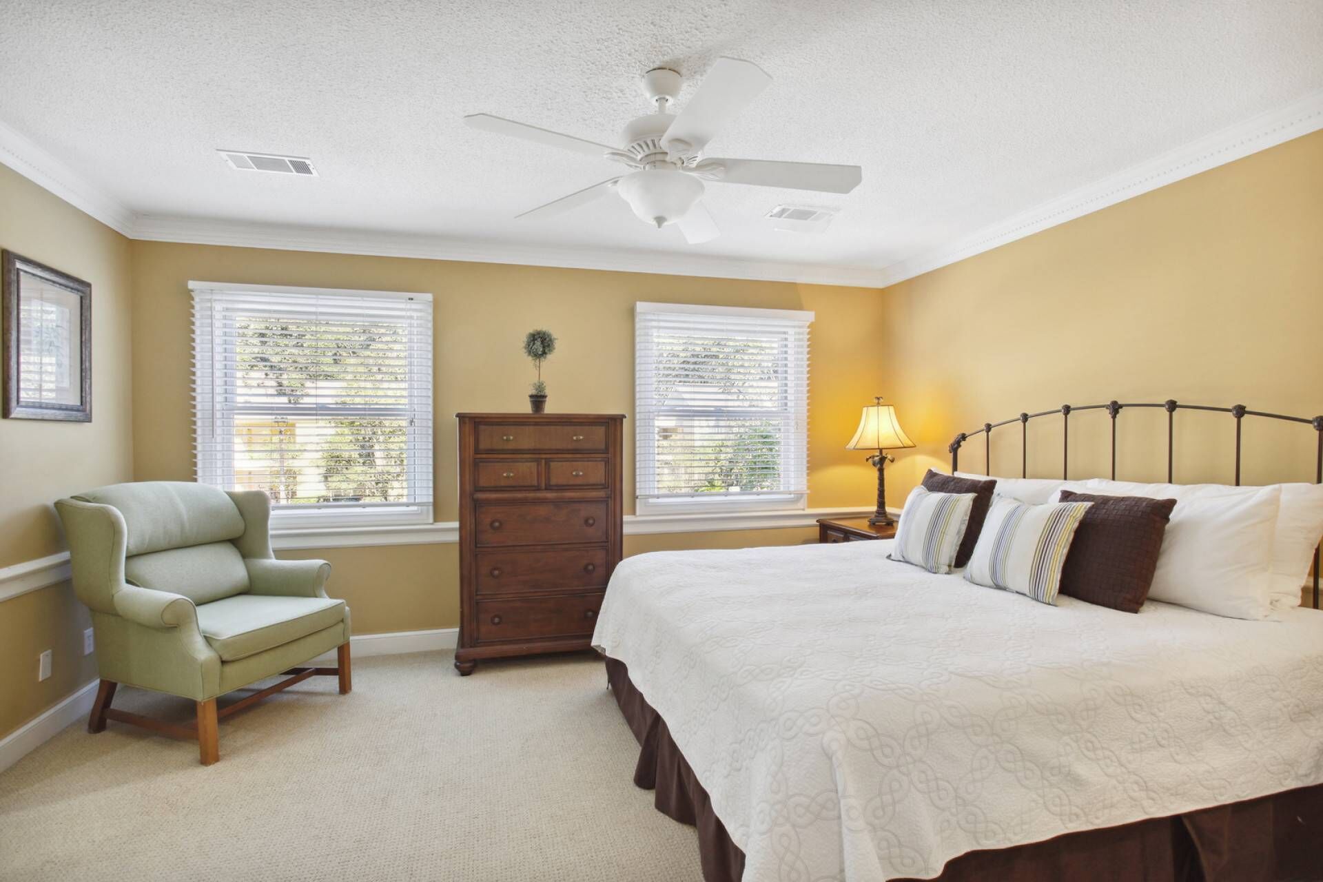 Une chambre à coucher comprenant un lit, une chaise, une commode et deux fenêtres équipées de stores.