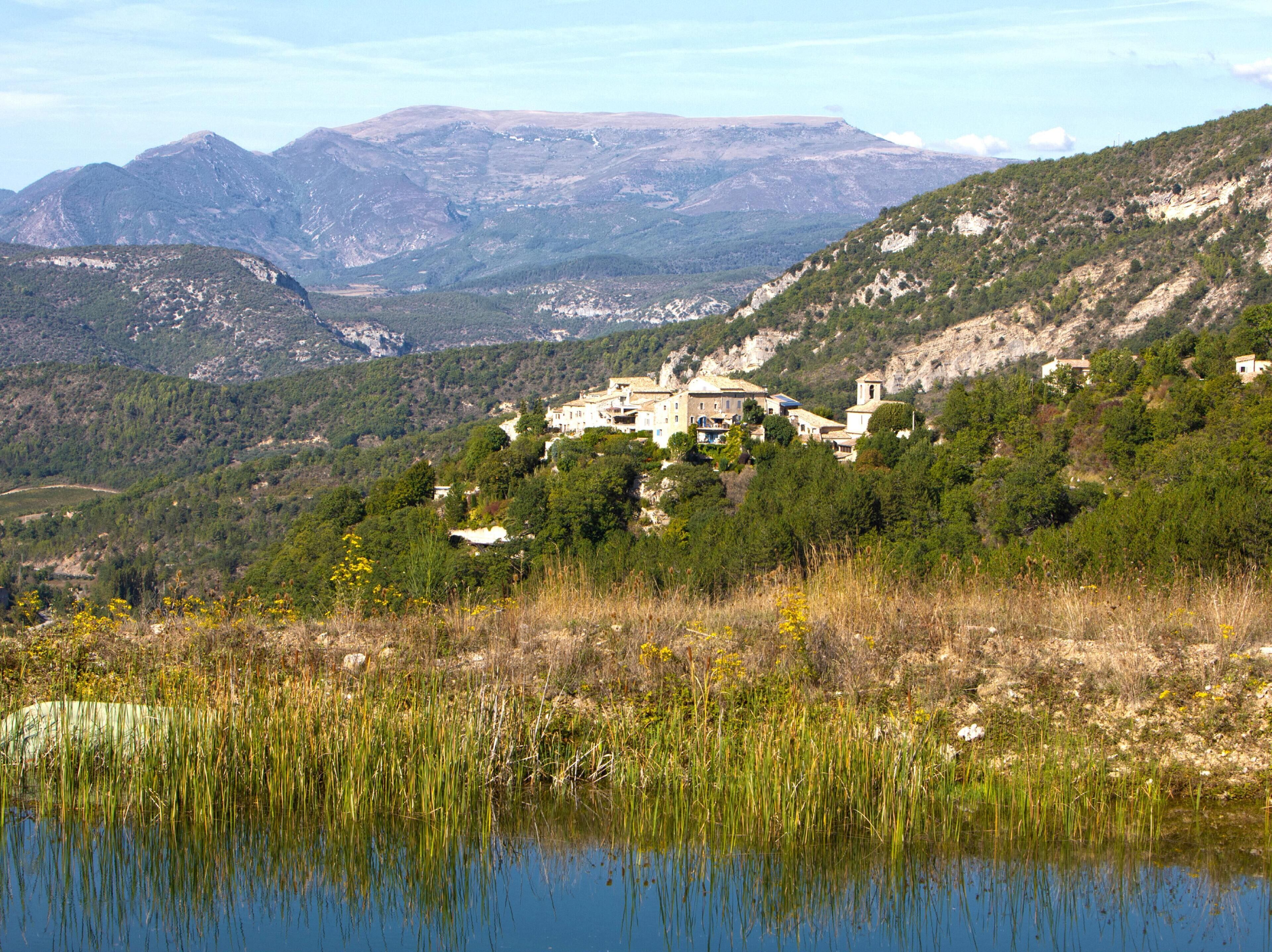 Paesaggio montuoso con un gruppo di edifici circondati dal verde e una distesa d'acqua in primo piano.