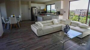A modern living room with a large L-shaped sofa, a dining area with a table and chairs, a kitchen with a central island, and large windows offering a city view. - Blue Luxury La Veleta (Tulum)