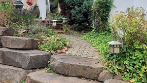 Un sendero de piedra que lleva a un jardín con valla blanca, una pérgola y una mesa con sillas.