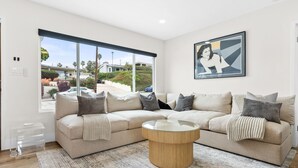 A modern living room with a beige sectional sofa, a glass coffee table, and a large window offering a view of the neighborhood.