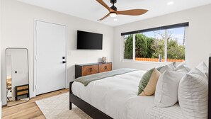 A bedroom with a bed, a ceiling fan, a TV mounted on the wall, and a window with a view of trees.