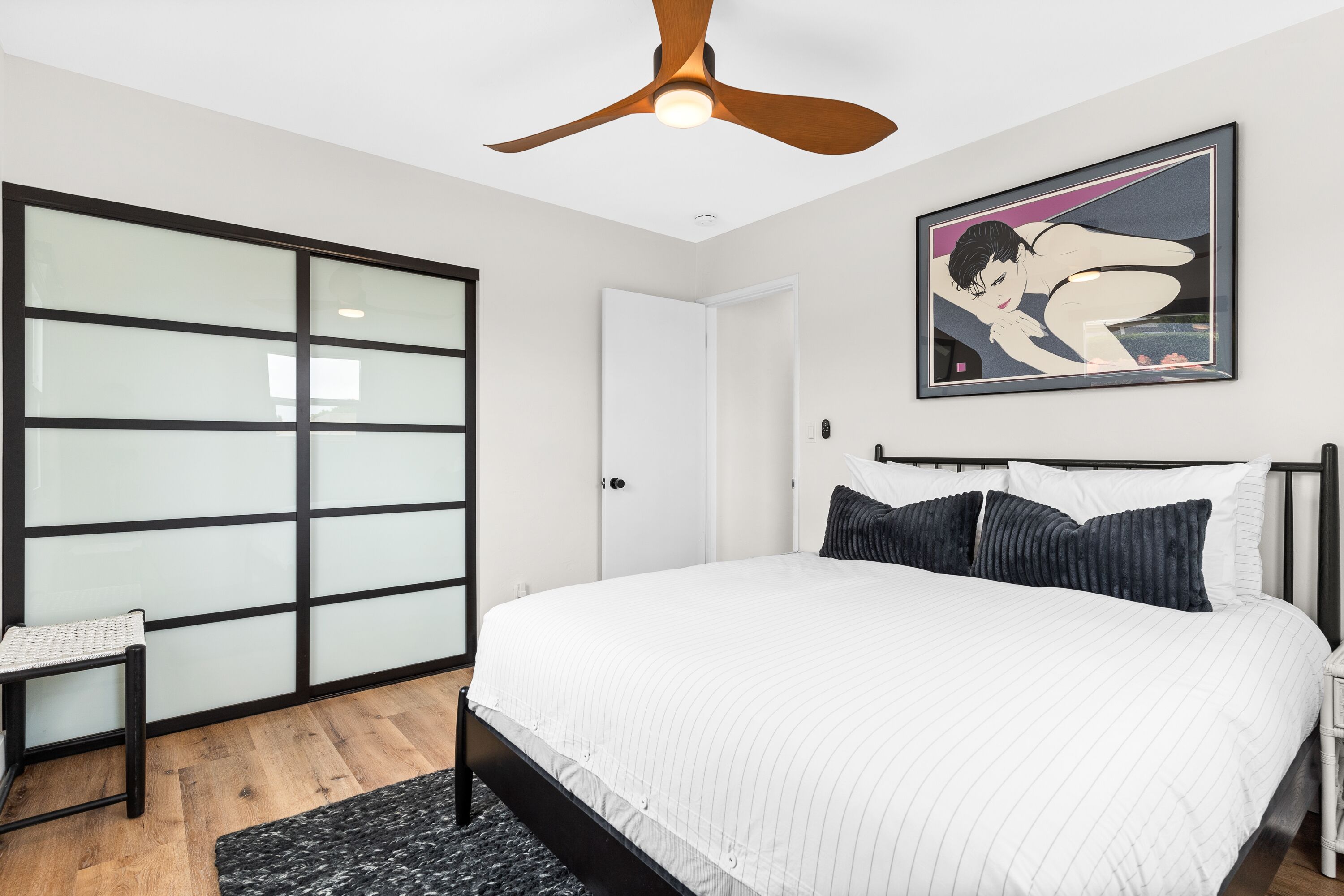 Ein Schlafzimmer mit einem Bett, einem Deckenventilator, einem gerahmten Bild, einer Schiebetür und einem kleinen Tisch.