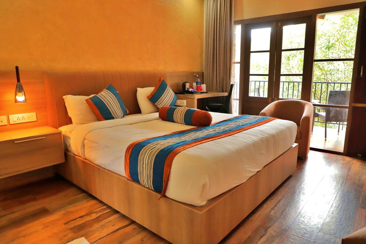 Habitación de hotel con una cama grande, cabecero de madera, un escritorio con una silla, una lámpara y vistas al exterior a través de una puerta corredera.