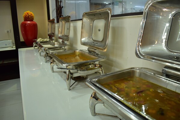 A buffet setup with multiple stainless steel chafing dishes containing various dishes, a red vase, and a window in the background. - Adrak Summer Sand Hill Resort Vagamon (Peermade)
