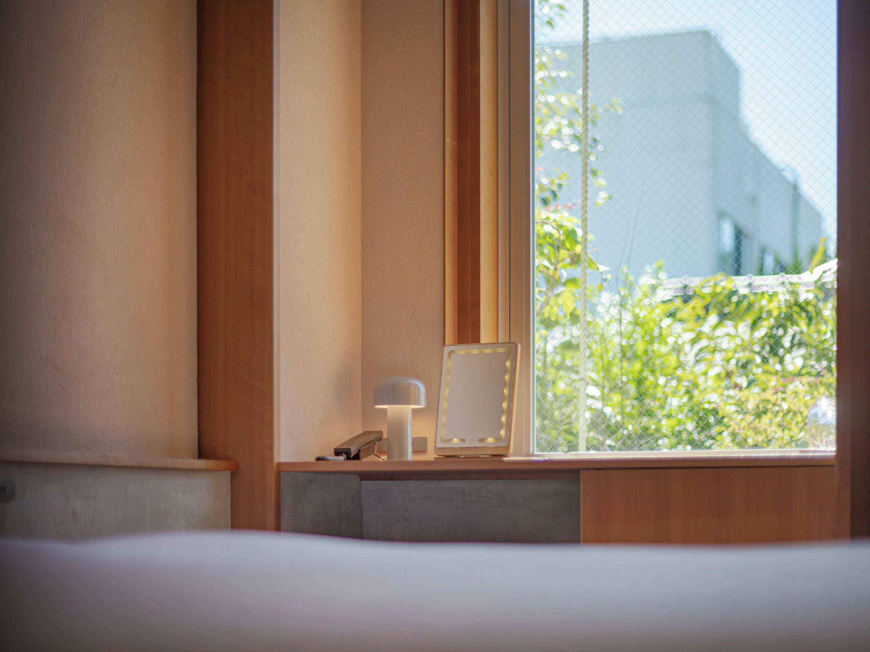 A hotel room with a bed, a bedside table with a lamp, a mirror, and a window with a view of greenery and a building.