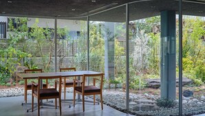 A modern dining area with a glass partition, wooden table, and chairs, overlooking a garden with various plants and a stone pathway.