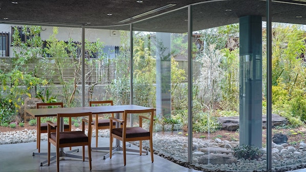 Et moderne spiseområde med en glasvæg, et træbord og stole, med udsigt til en have med forskellige planter og en stensti.