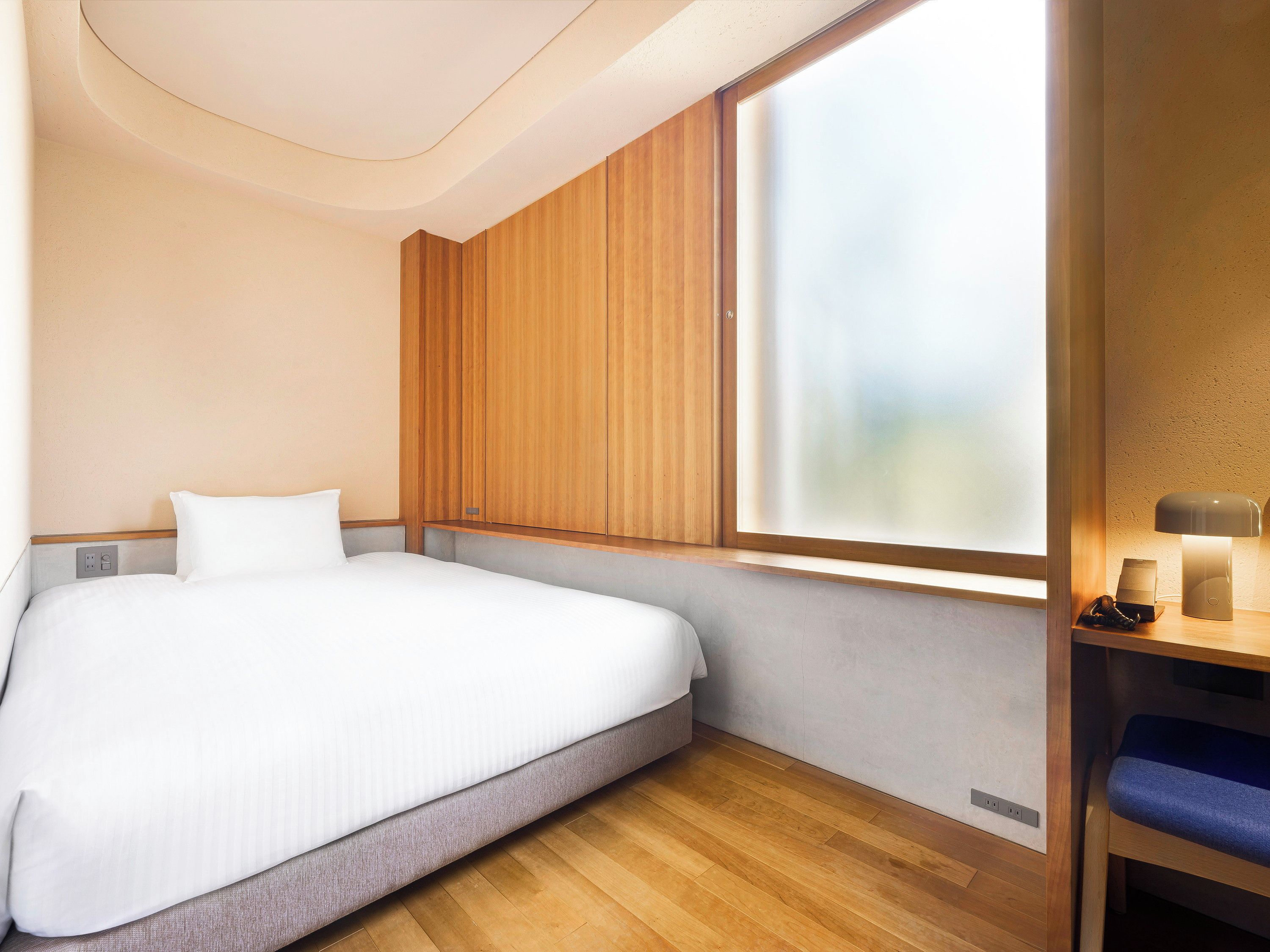 A modern hotel room with a bed, a window, a bedside table with a lamp, and a small shelf.