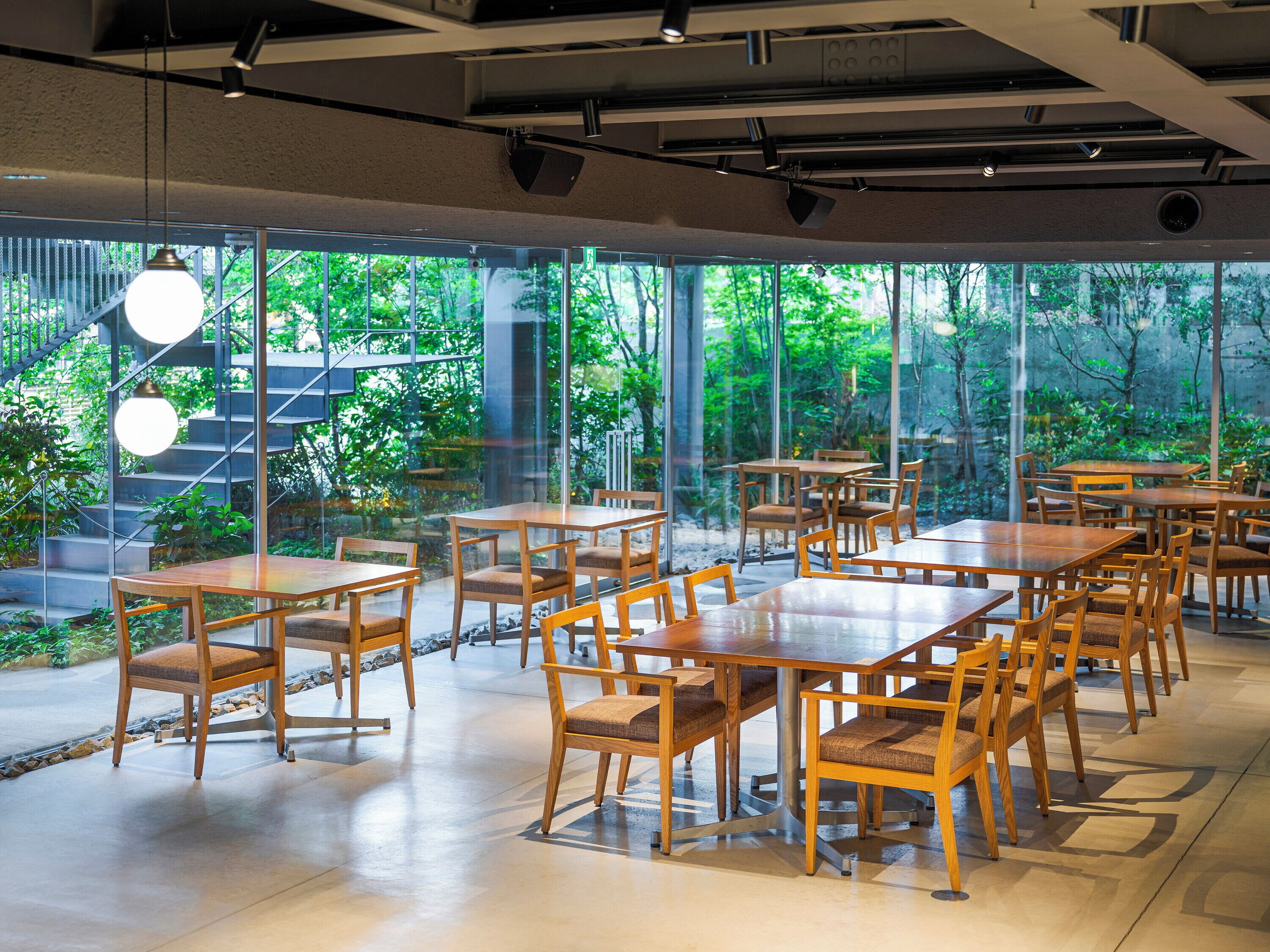 A restaurant with wooden tables and chairs, large windows, and a view of greenery.