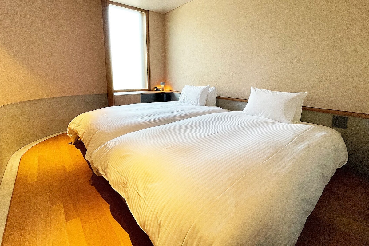 Una habitación de hotel con dos camas, una mesita de noche y una ventana.