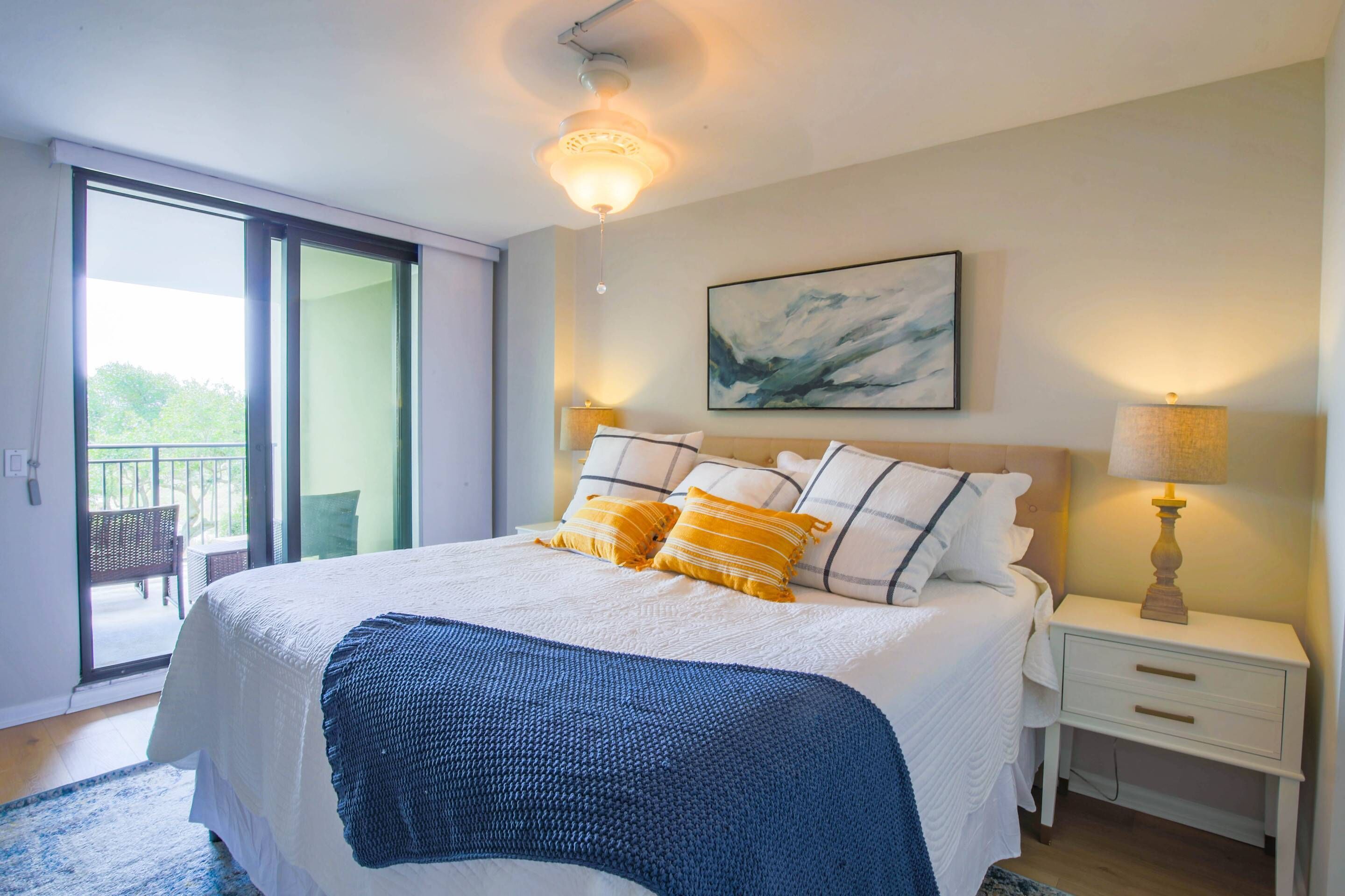 A bedroom with a large bed, a nightstand with a lamp, a painting on the wall, and a sliding glass door leading to an outdoor area.