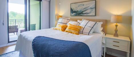 A bedroom with a large bed, a nightstand with a lamp, a painting on the wall, and a sliding glass door leading to an outdoor area.