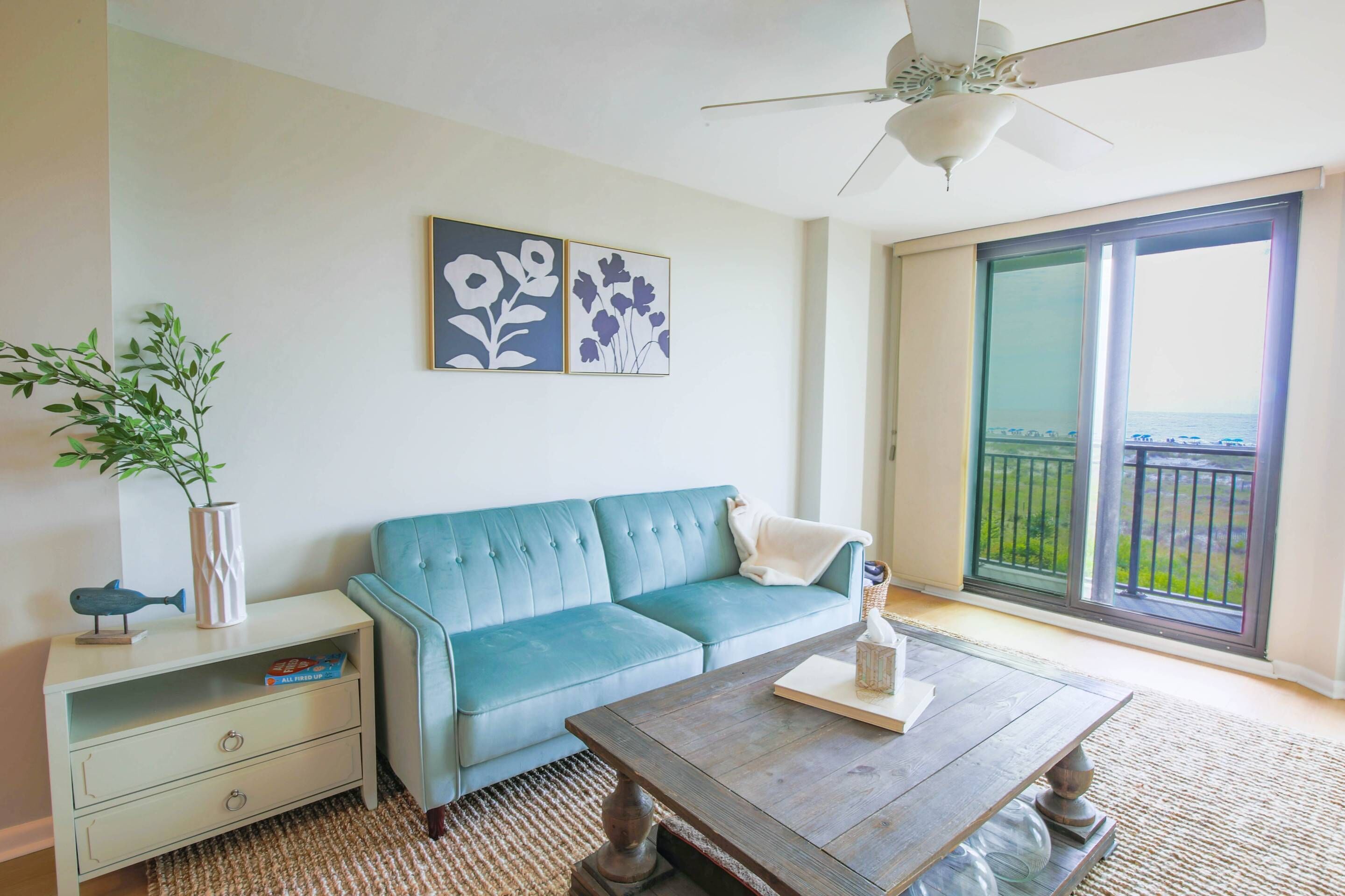 A living room with a blue sofa, a wooden coffee table, a white side table, and a large window with a view of the outdoors.