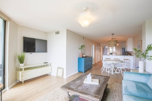 A modern living room with a flat-screen TV, a coffee table, and a sofa. There is a kitchen area in the background with a dining table and chairs.