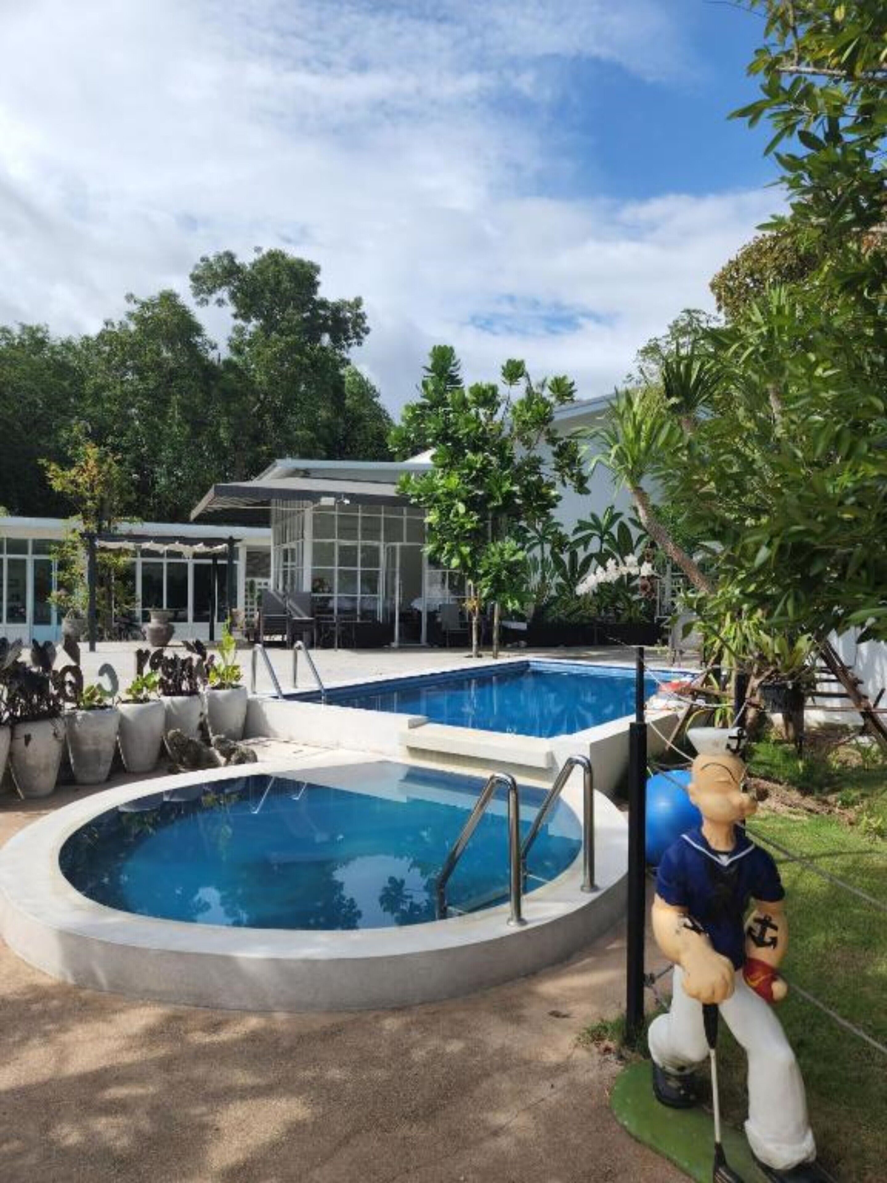 Zona de piscina con una estatua de un marinero, rodeada de plantas en macetas y un edificio al fondo.