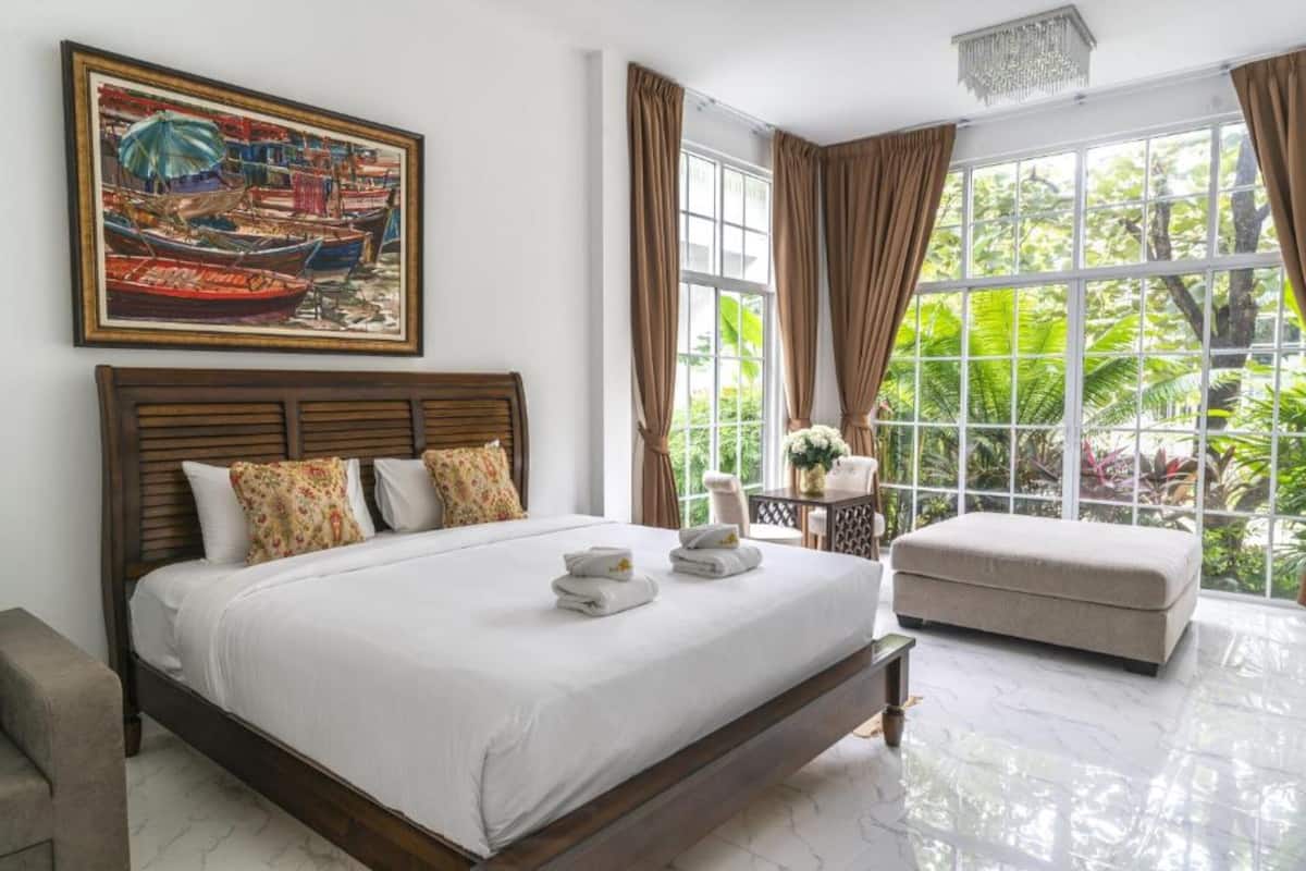 Une chambre d’hôtel avec un grand lit, une petite table sur laquelle est posé un vase, et une vue sur la verdure grâce à de grandes fenêtres.
