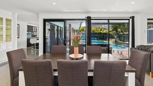 Un espace repas moderne avec une table et des chaises, un vase de fleurs et une vue sur une piscine, visible à travers de grandes portes vitrées.
