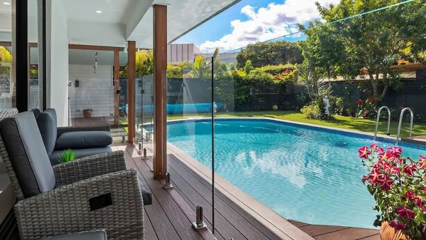Un espace piscine extérieur moderne, doté de rampes en verre, de meubles en osier et offrant une vue sur un jardin à l’arrière de la maison.