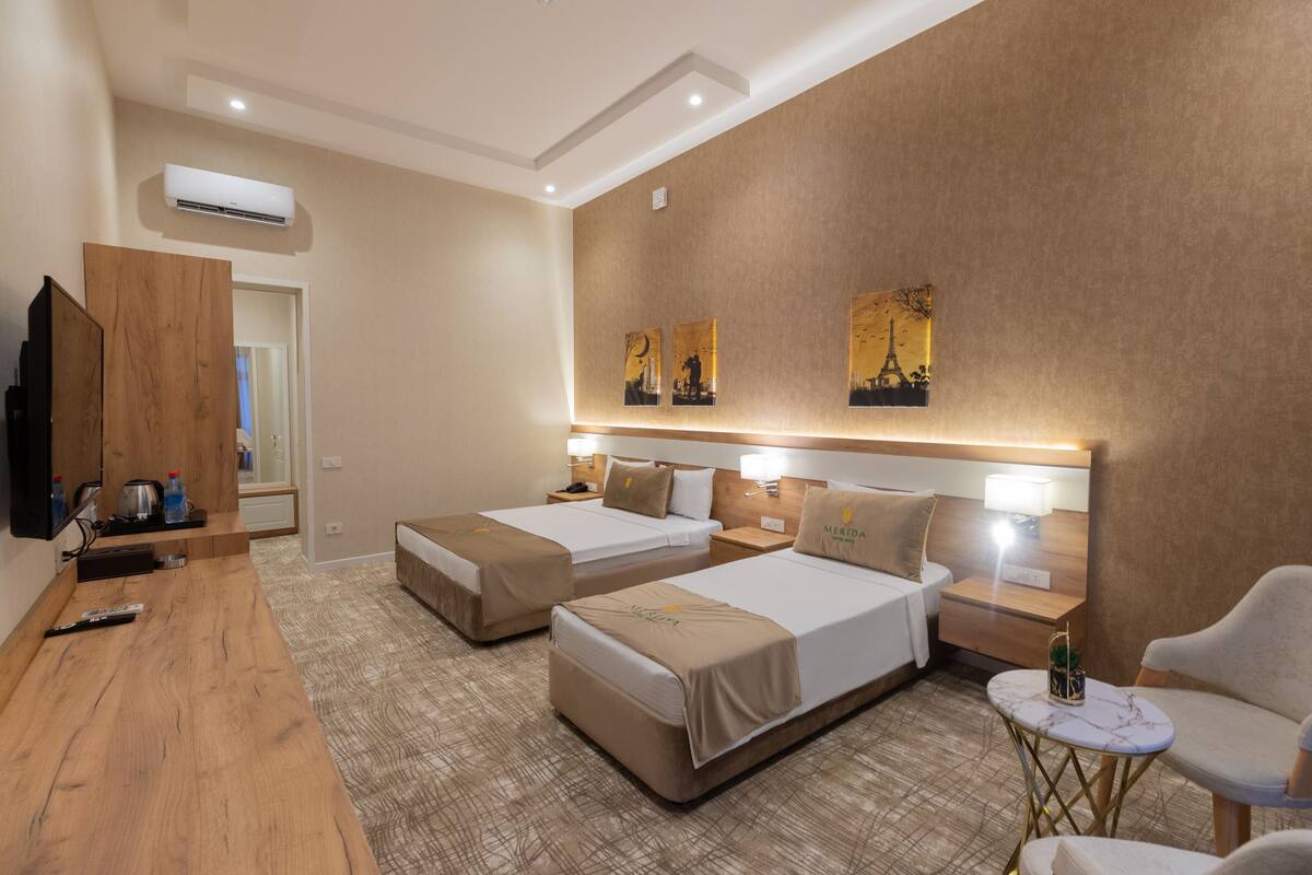 A hotel room with two beds, a TV, a wooden desk, a small table, and artwork on the wall.