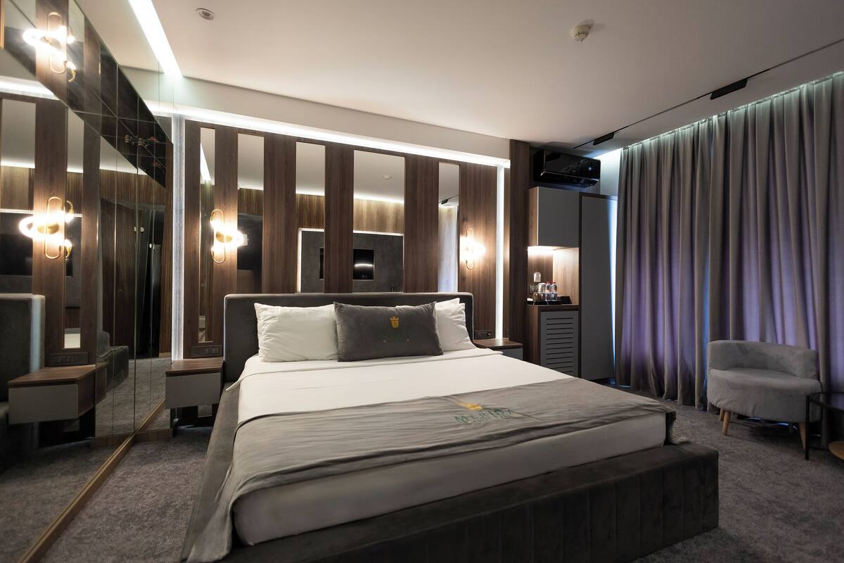 A modern hotel room with a large bed, a bedside table, a chair, a mirror, and a curtain.