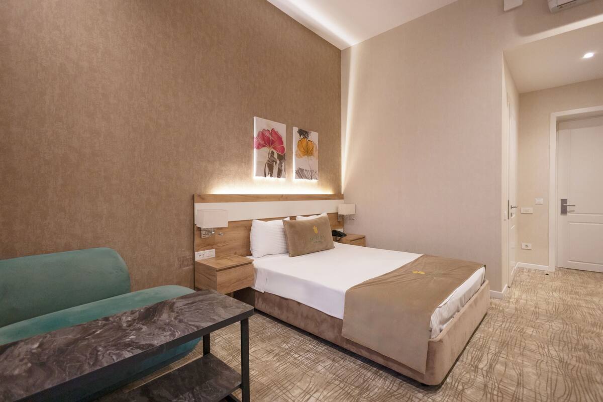 A hotel room with a bed, a nightstand, a table, a sofa, and two wall-mounted artworks.