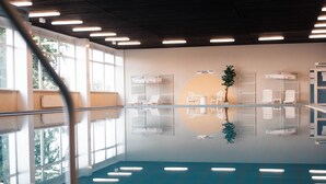 Piscina interior com paredes de vidro transparente, uma planta central e espreguiçadeiras.