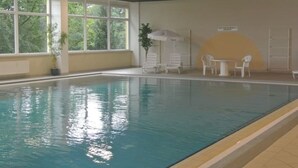 Una piscina interior con agua cristalina, ventanas amplias y algunas sillas y mesas.