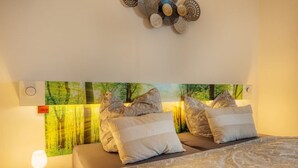 Un dormitorio con cama, almohadas, mesita de noche, lámpara y una obra de arte con árboles.