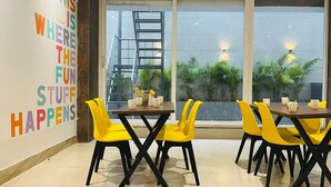 Un espacio interior moderno con zona de comedor, sillas amarillas y una mesa de madera, ventanas amplias con vistas a zonas verdes y una cita motivacional en la pared.