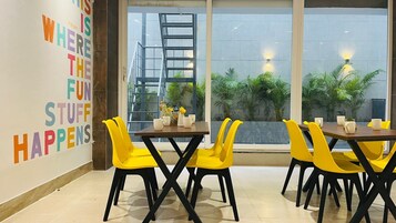 A modern indoor space with a dining area featuring yellow chairs and a wooden table, large windows with a view of greenery, and a motivational quote on the wall.