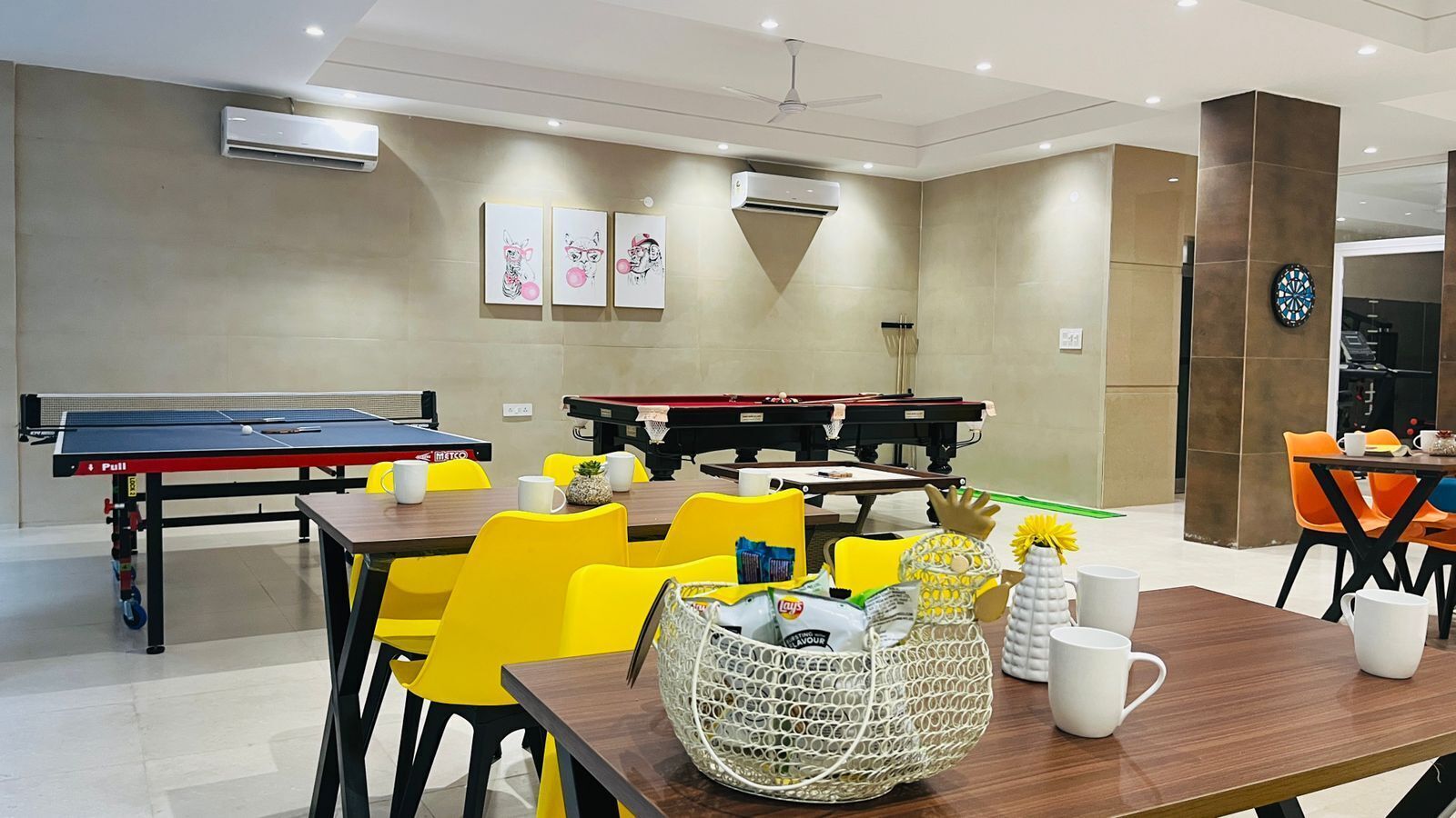 A room with ping pong tables, billiards tables, and yellow chairs. There are air conditioning units on the walls and a clock on the right side.
