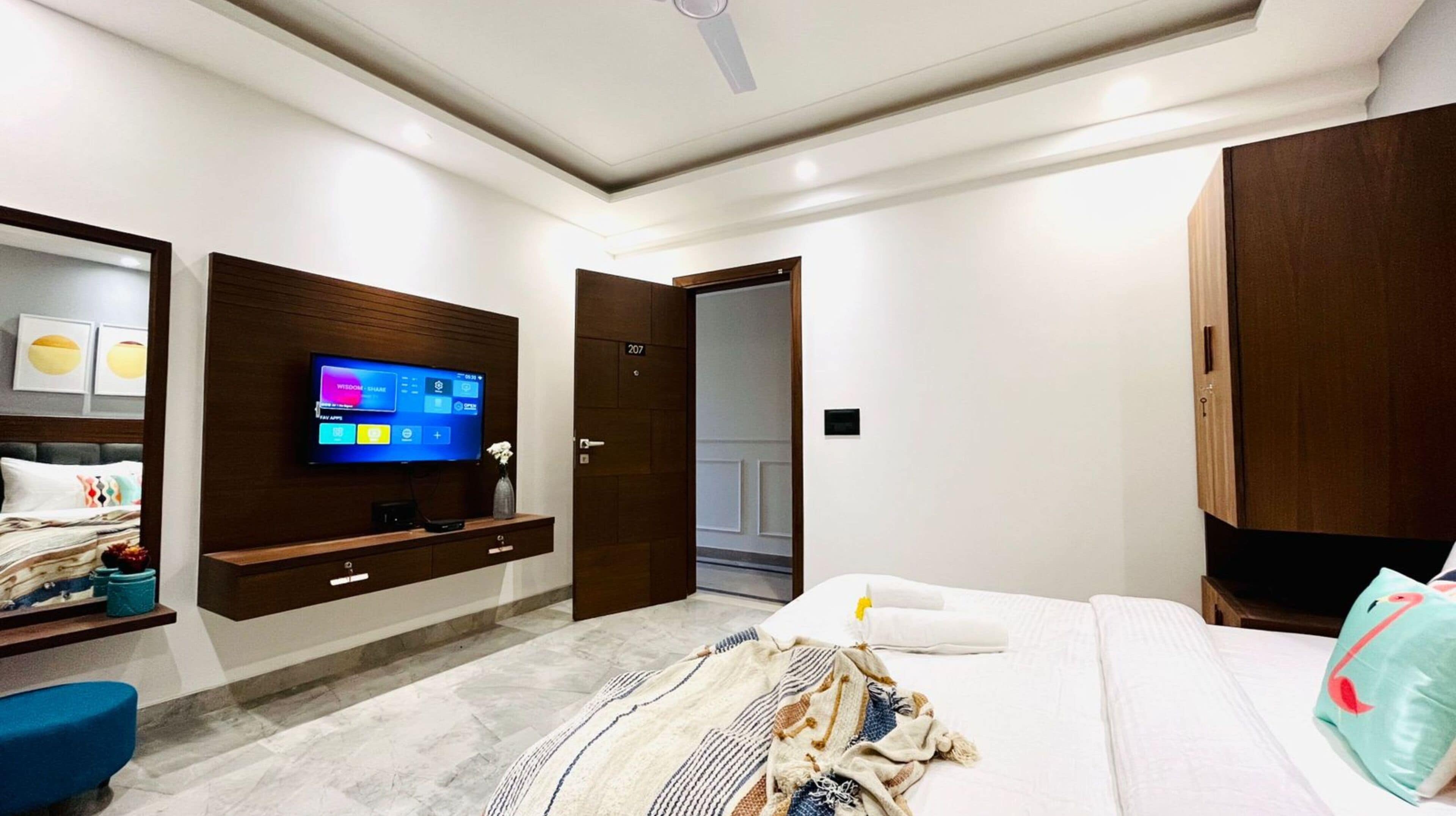 A modern hotel room with a large bed, a flat-screen TV mounted on the wall, a wooden wardrobe, and a small side table with a vase.